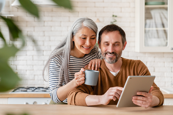 Two middle-aged adults looking at a tablet computer in a kitchen. Female subject leans on shoulder of male subject while holding a coffee mug.